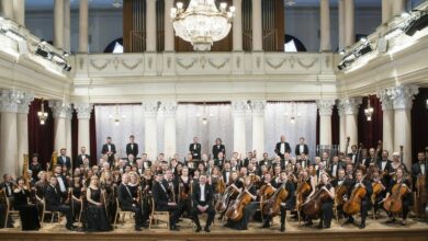 Das Nationale Symphonieorchester der Ukraine posiert 2018 mit seinen Instrumenten auf der Bühne. Sie tragen eine formelle schwarze Krawatte und das Gebäude ist mit einem großen Glaskronleuchter über ihnen verziert.