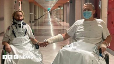 Saule Linkus, in a neck brace, holds hands with husband Darius Linkus as the pair sit in wheelchairs in a hospital corridor