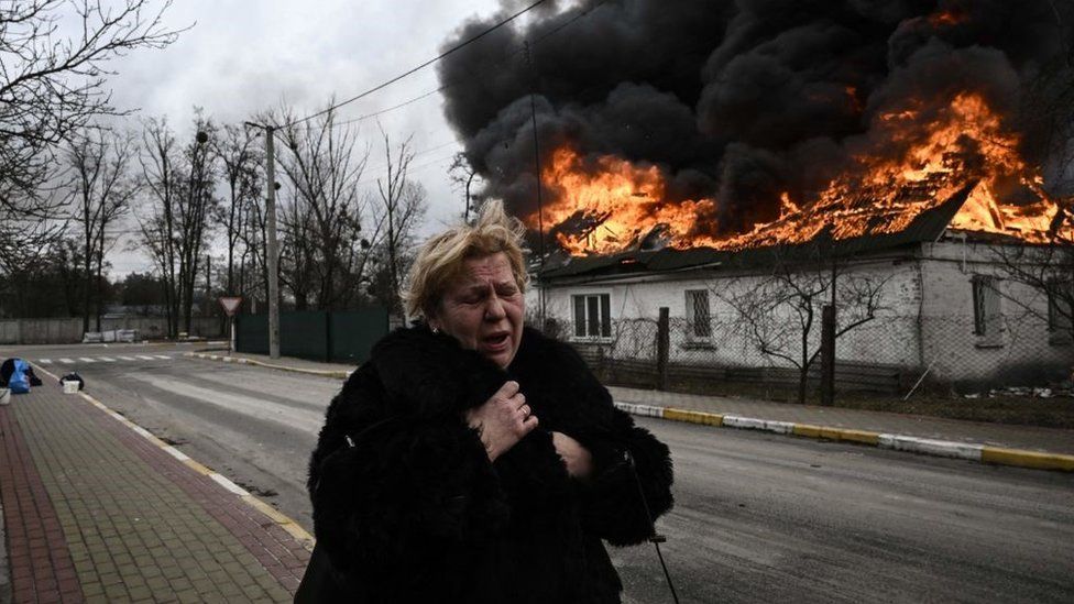Eine Frau steht im März 2022 in der Stadt Irpin außerhalb von Kiew vor einem Haus, das nach einem Beschuss brennt